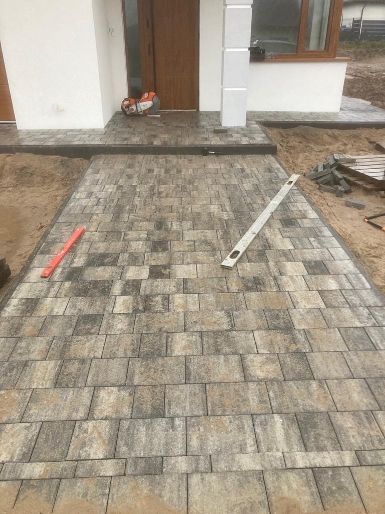 Świeżo ułożona kostka brukowa w odcieniach szarości i beżu prowadzi do wejścia domu. Widoczne narzędzia: poziomica i piła do cięcia kostki. Częściowo zasypane piaskiem obrzeża.