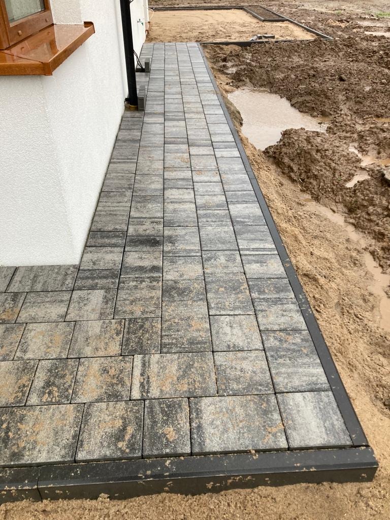 Świeżo ułożony chodnik z szarej kostki brukowej z czarną obwódką, prowadzący wzdłuż białej ściany budynku, z widocznym błotnistym terenem w tle.