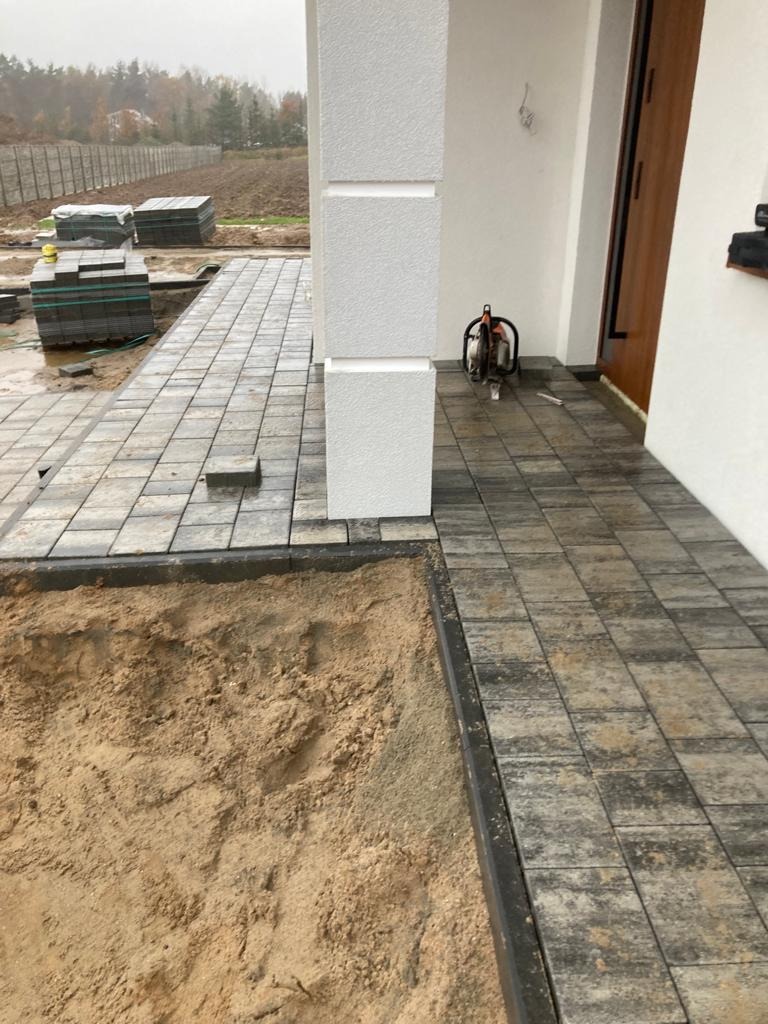 Częściowo ułożona ścieżka z kostki brukowej w odcieniach szarości, obok przygotowane podłoże z piasku, palety kostki i przecinarka do kostki brukowej przy ścianie domu.