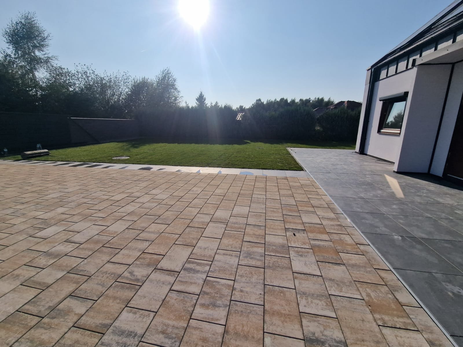 Beżowo-brązowa kostka brukowa ułożona na podjeździe przed nowoczesnym domem z trawnikiem w tle, widok w słoneczny dzień.