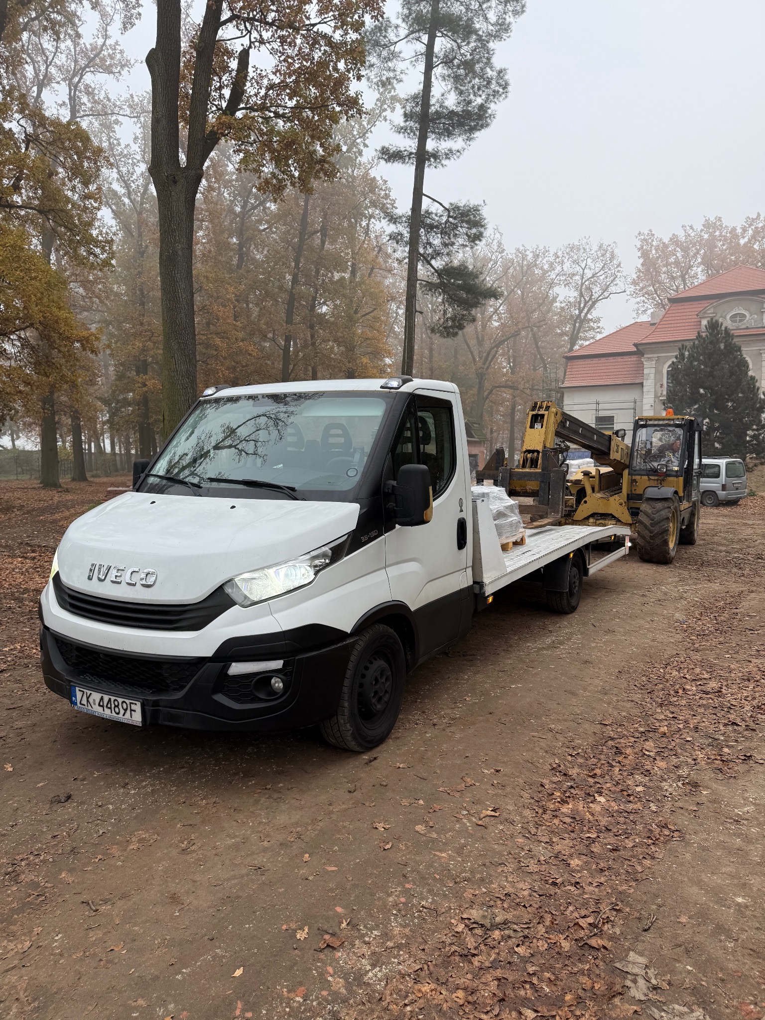 Biały samochód transportowy Iveco z żółtą maszyną budowlaną na platformie, zaparkowany na ziemistej drodze pokrytej liśćmi, w tle drzewa i budynek z czerwoną dachówką w mglisty dzień.
