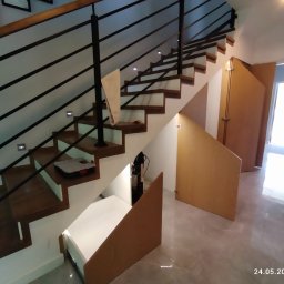 Oksana Shulyar Firma Produkcyjno-Usługowa VIDA MEBBUD - Schody drewniane z czarną metalową balustradą i zabudową wnęki pod schodami z jasnego drewna, widoczne otwarte schowki z białymi półkami.