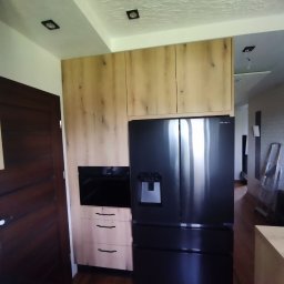 Oksana Shulyar Firma Produkcyjno-Usługowa VIDA MEBBUD - Nowoczesna zabudowa kuchenna z drewnianymi frontami, czarnym piekarnikiem i dużą lodówką side-by-side z dystrybutorem wody, widok z perspektywy podłogi, oświetlenie sufitowe punktowe.