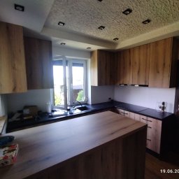 Oksana Shulyar Firma Produkcyjno-Usługowa VIDA MEBBUD - Nowoczesna kuchnia z drewnianymi frontami i wyspą, czarny blat, okno z widokiem na zieleń, dekoracyjny sufit z oświetleniem punktowym.