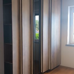 Oksana Shulyar Firma Produkcyjno-Usługowa VIDA MEBBUD - Wysoka, drewniana szafa z oświetleniem LED w pionowych listwach, z otwartą wnęką z lustrem, widok na zewnątrz przez okno.