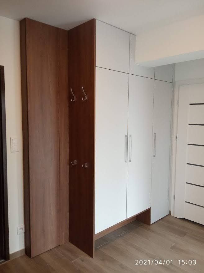 Szafa wnękowa z białymi frontami i drewnianym wykończeniem, z otwartą częścią z wieszakami, dopasowana do kąta w przedpokoju.