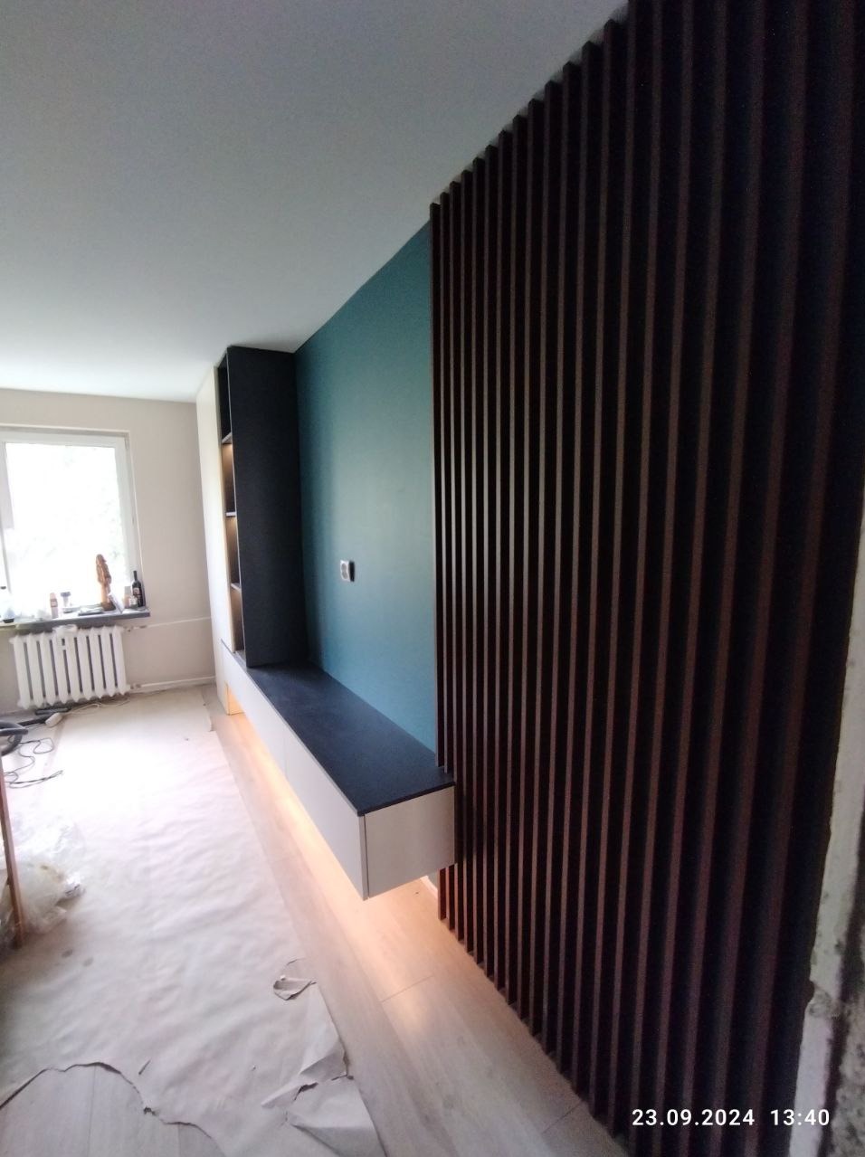 Nowoczesna ściana z drewnianych lameli w kolorze ciemnego brązu przylegająca do ściany w kolorze morskim. Na ścianie zamontowana podświetlana szafka z półkami. W tle okno i grzejnik.