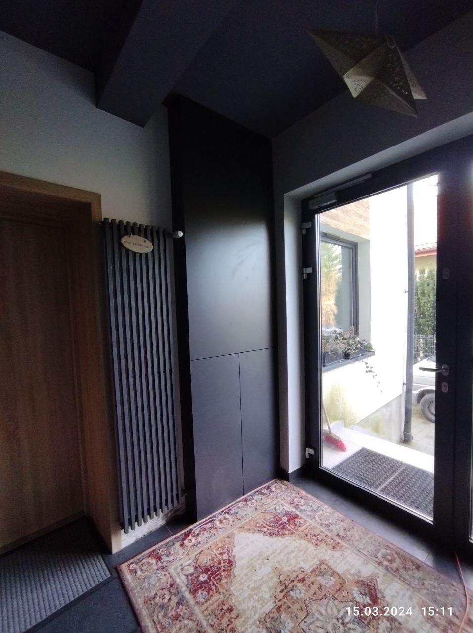 Wnętrze przedpokoju z ciemną zabudową meblową, grzejnikiem panelowym i wzorzystym dywanem, widok na zewnątrz przez przeszklone drzwi wejściowe.