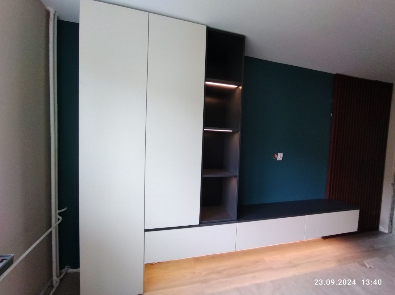 Biało-szara zabudowa meblowa z oświetleniem LED w półkach, na tle ściany w kolorze butelkowej zieleni i drewnianych paneli dekoracyjnych.