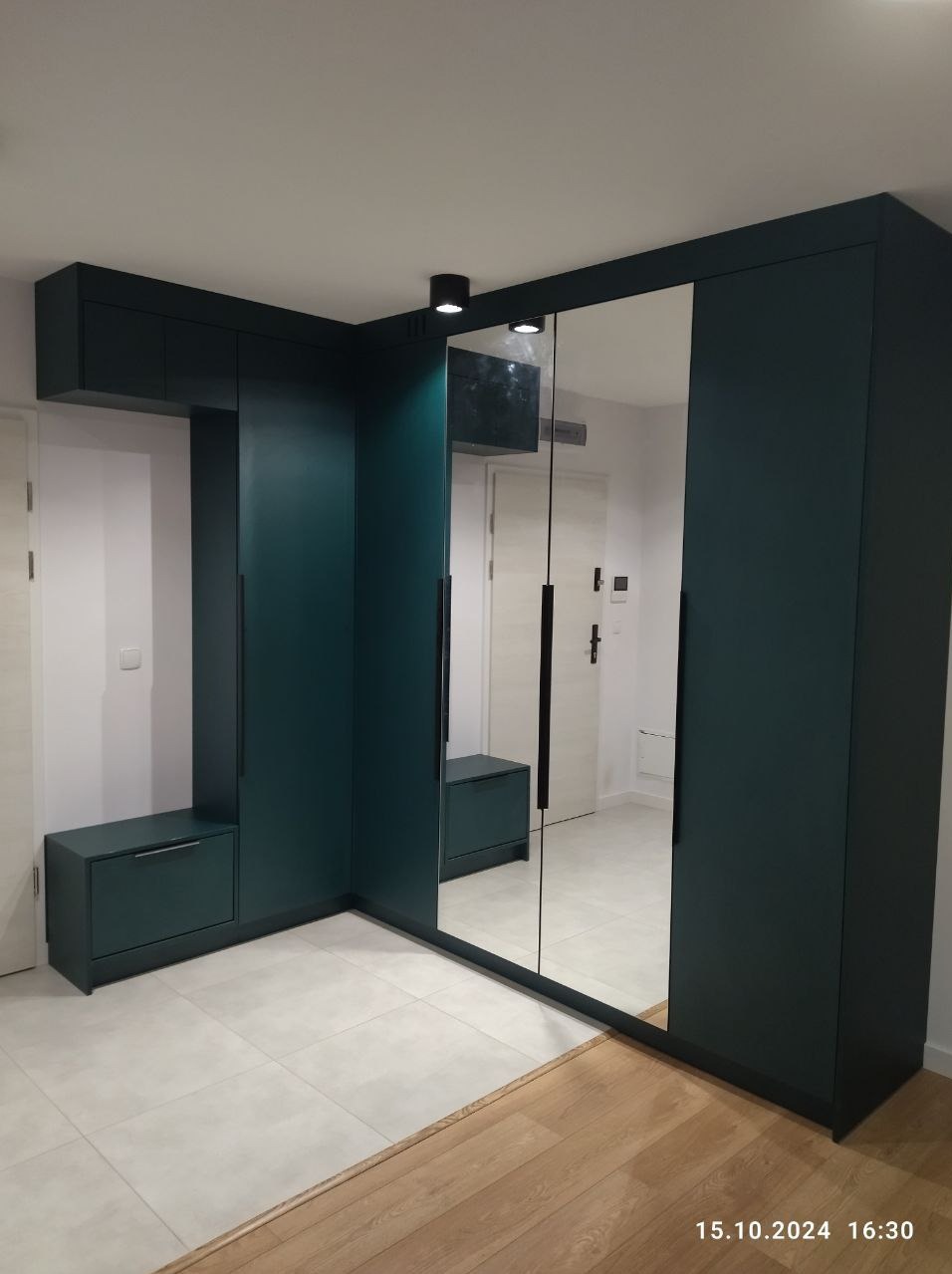 Ciemnozielona szafa wnękowa z lustrzanymi drzwiami, wbudowana w narożnik przedpokoju, z dodatkowymi szafkami i siedziskiem, oświetlenie punktowe.