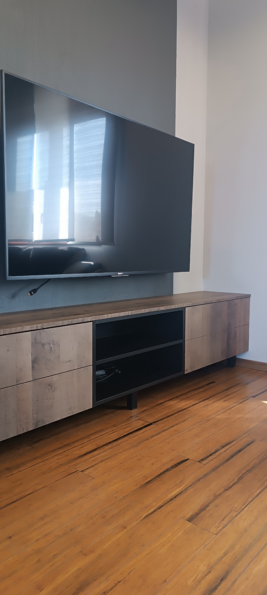 Nowoczesna szafka RTV z drewnianymi frontami i otwartą przestrzenią na sprzęt, wiszący telewizor na szarej ścianie, drewniana podłoga.