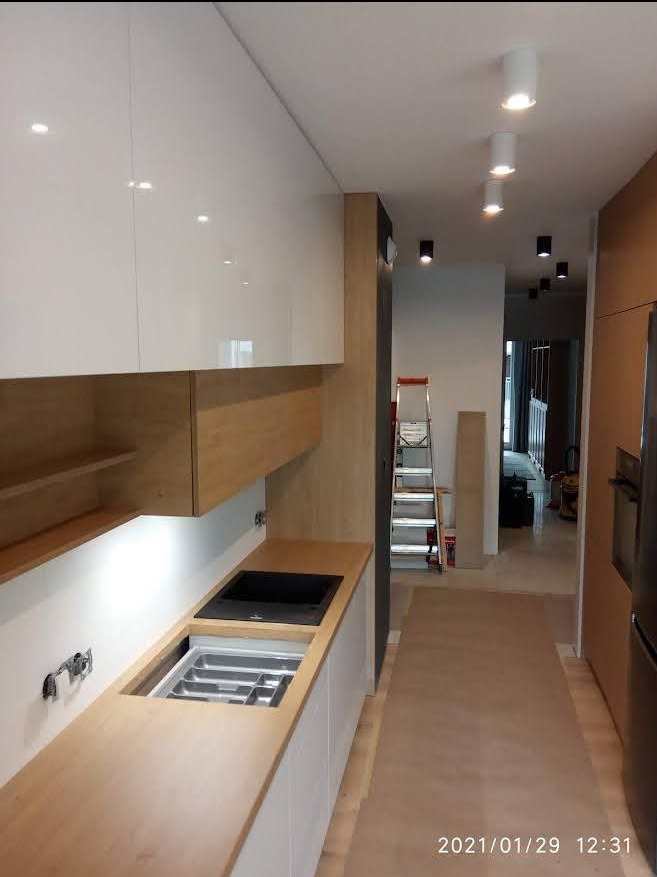 Montaż nowoczesnej kuchni: widoczne białe, błyszczące szafki górne, drewniane szafki dolne z wbudowanym zlewem i ociekaczem, oraz drabina w tle, sugerująca trwający proces instalacji.