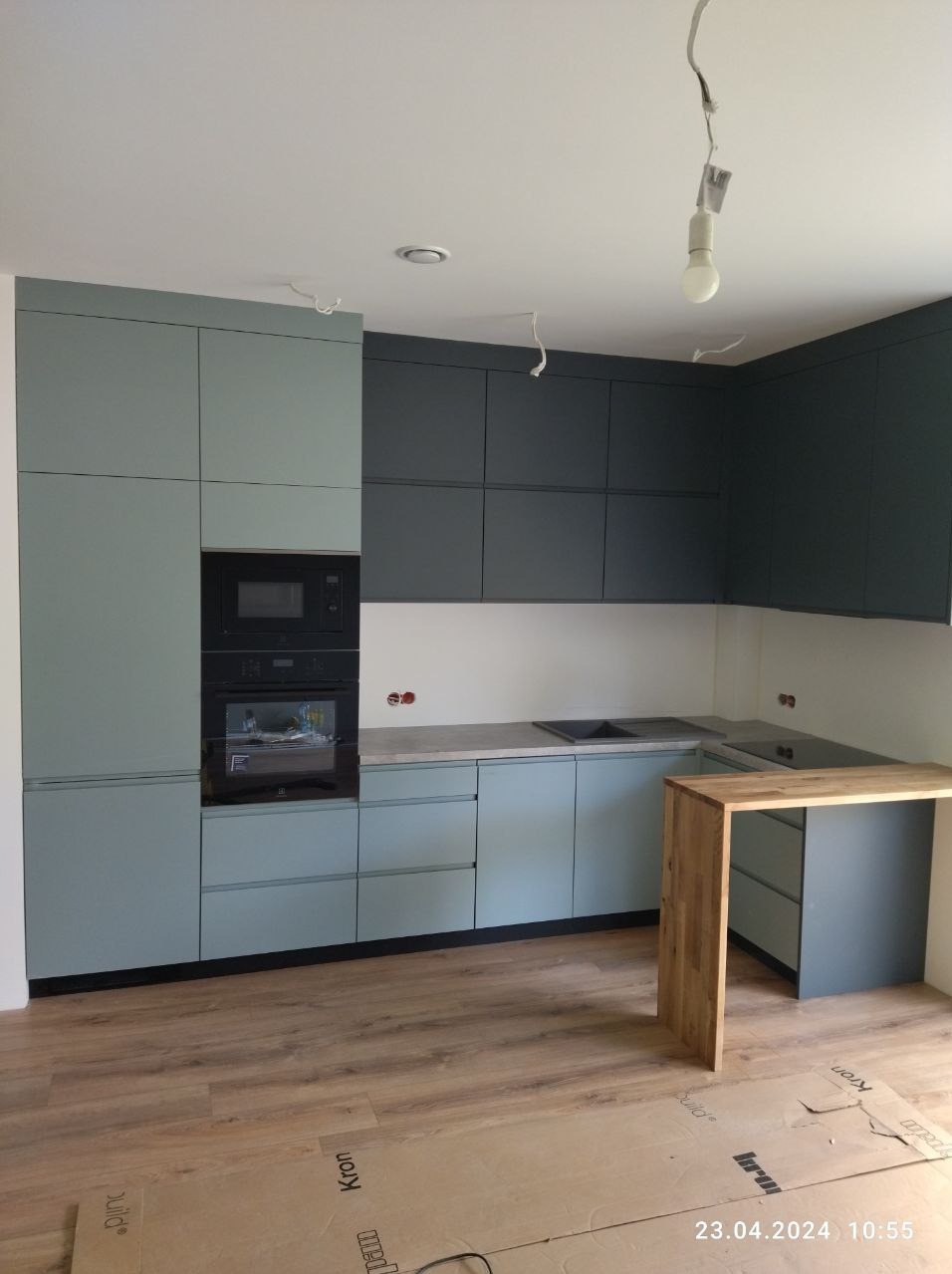 Nowoczesna kuchnia z zabudową w odcieniach szarości i błękitu, z drewnianym blatem-wyspą i widocznymi przewodami elektrycznymi przed finalnym wykończeniem.