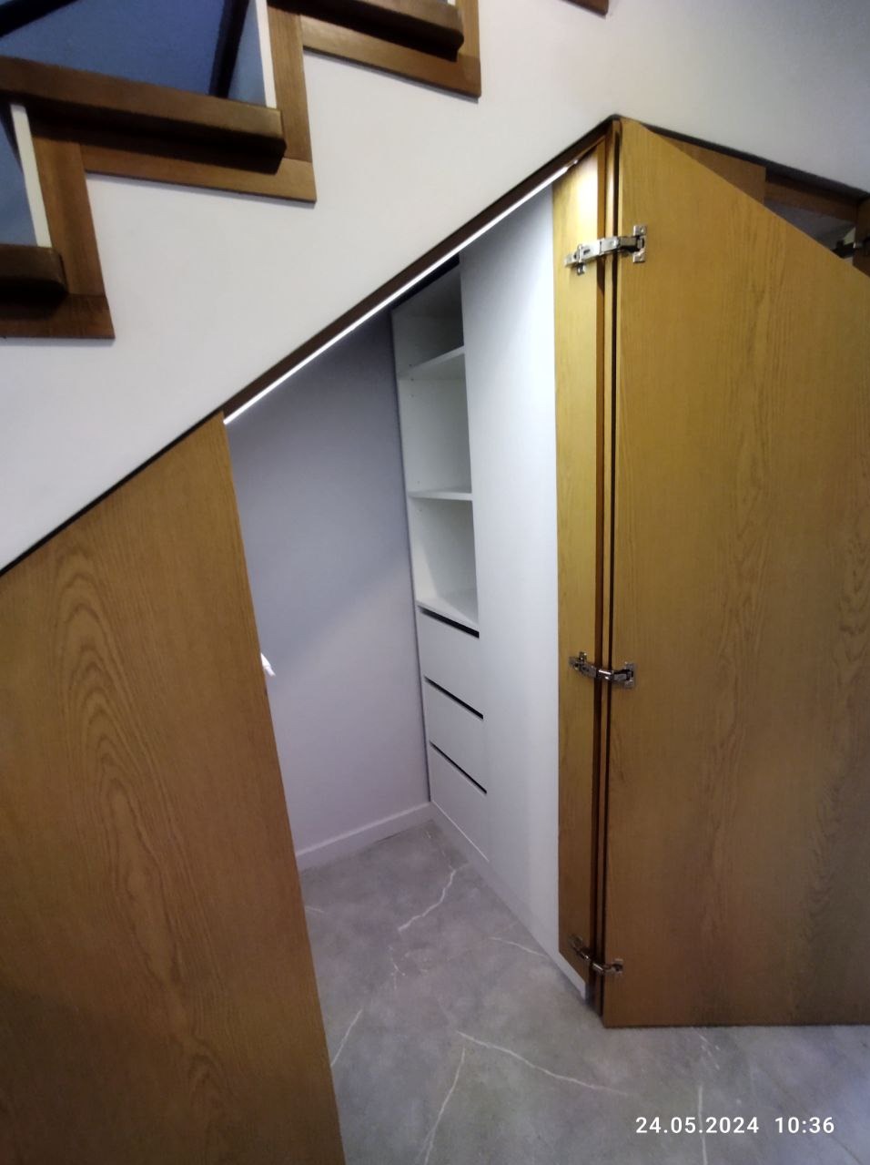 Szafa wnękowa z drewnianymi drzwiami pod schodami, z widocznymi półkami i szufladami wewnątrz.