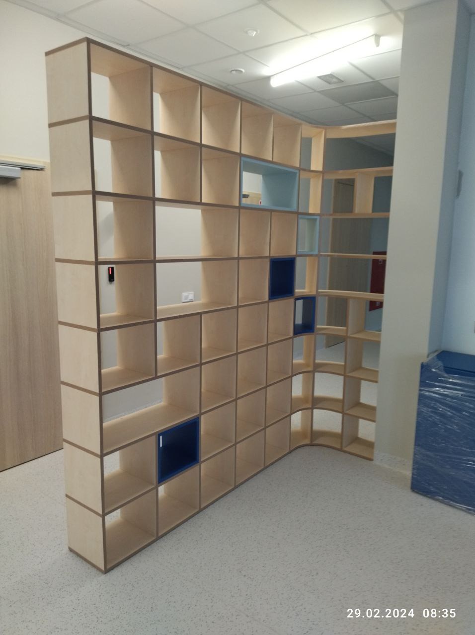 Duża, drewniana, ażurowa szafa na wymiar z elementami w kolorze niebieskim, pełniąca funkcję ścianki działowej w jasnym pomieszczeniu.