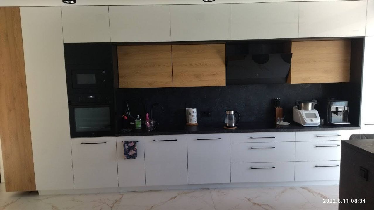 Nowoczesna kuchnia z białymi szafkami dolnymi i górnymi, drewnianymi akcentami, czarnym blatem i sprzętem AGD, z widocznym ręcznikiem z motywem kwiatowym na jednej z szafek.