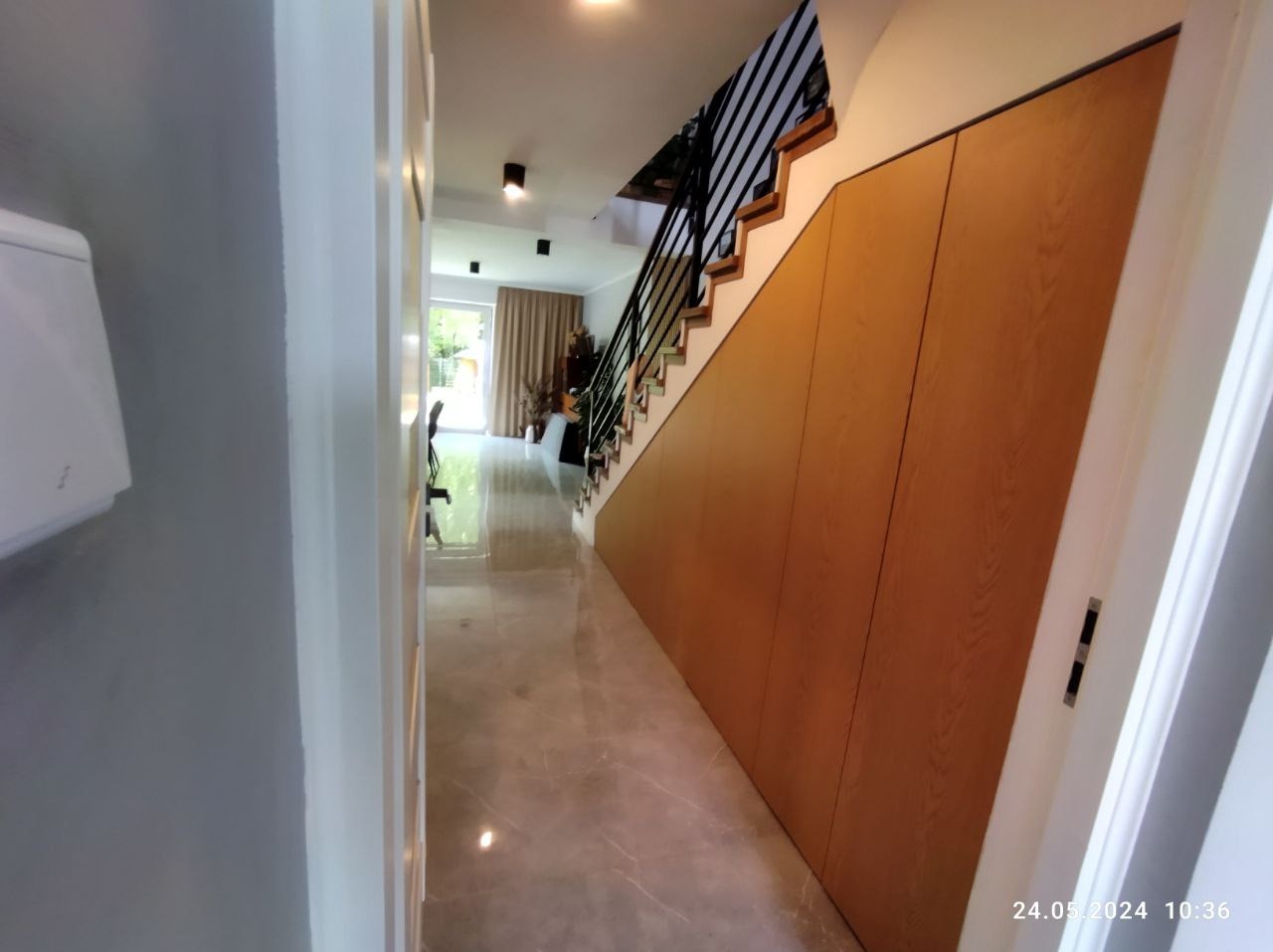 Wnętrze domu z szafą na wymiar pod schodami, drewniane fronty bez uchwytów, czarne metalowe schody, marmurowe płytki podłogowe.