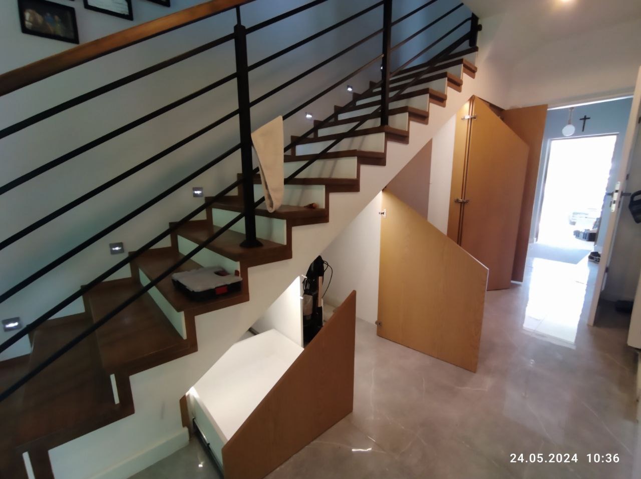 Schody drewniane z czarną metalową balustradą i zabudową wnęki pod schodami z jasnego drewna, widoczne otwarte schowki z białymi półkami.