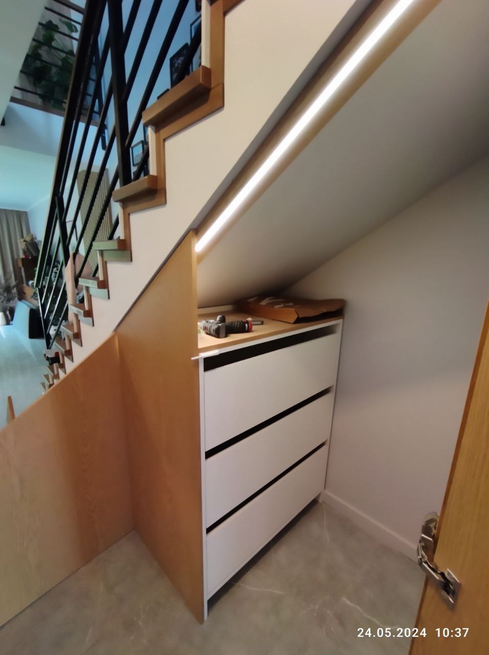 Biała komoda z trzema szufladami w przestrzeni pod schodami, z oświetleniem LED i narzędziami na blacie, w trakcie montażu obudowy schodów z płyty meblowej.