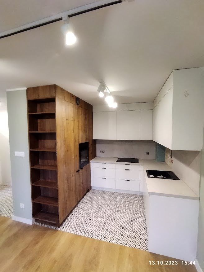 Nowoczesna kuchnia z białymi szafkami i drewnianą zabudową z piekarnikiem i regałem, oświetlenie punktowe na suficie, wzorzysta podłoga.