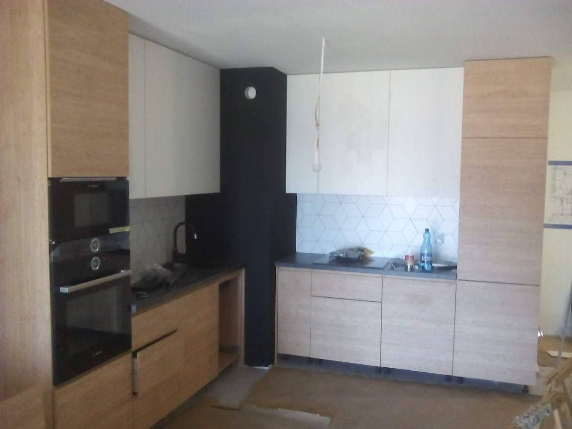 Montaż nowoczesnej kuchni: jasne, drewnopodobne fronty szafek dolnych i górnych kontrastują z białymi szafkami wiszącymi i czarną ścianą. Widoczny piekarnik do zabudowy i fragment mozaiki na ścianie.