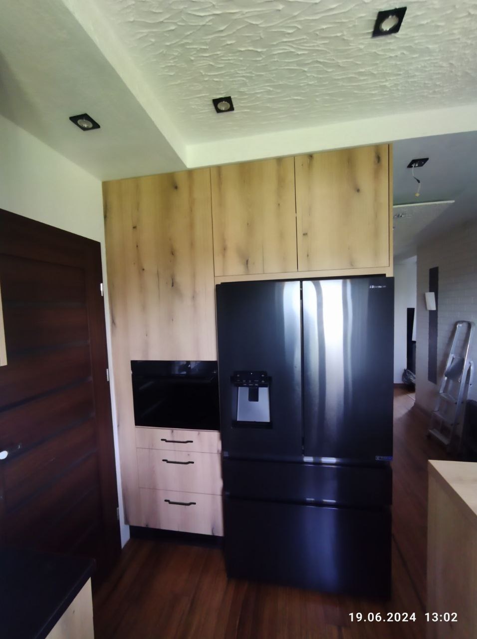 Nowoczesna zabudowa kuchenna z drewnianymi frontami, czarnym piekarnikiem i dużą lodówką side-by-side z dystrybutorem wody, widok z perspektywy podłogi, oświetlenie sufitowe punktowe.