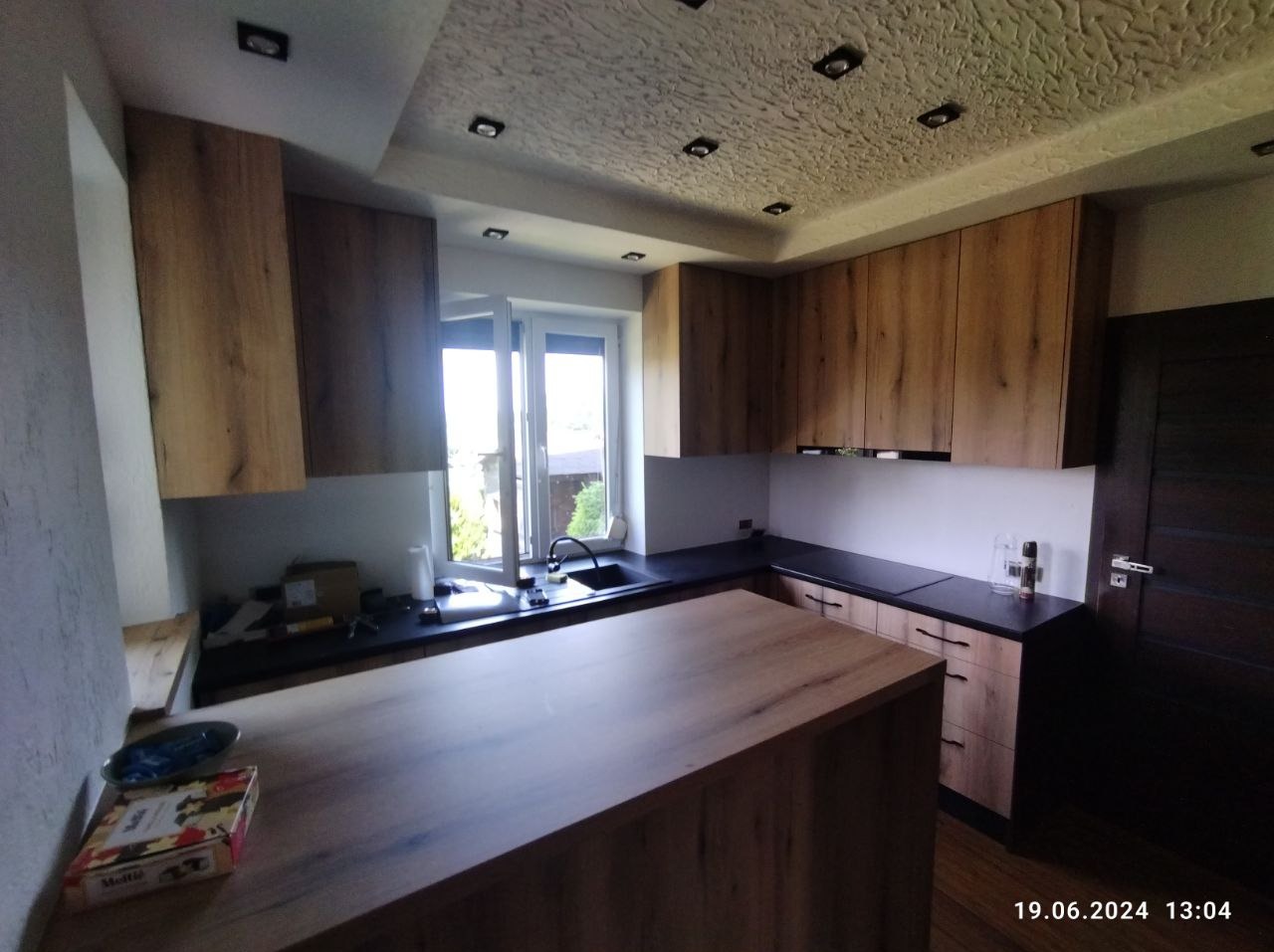 Nowoczesna kuchnia z drewnianymi frontami i wyspą, czarny blat, okno z widokiem na zieleń, dekoracyjny sufit z oświetleniem punktowym.