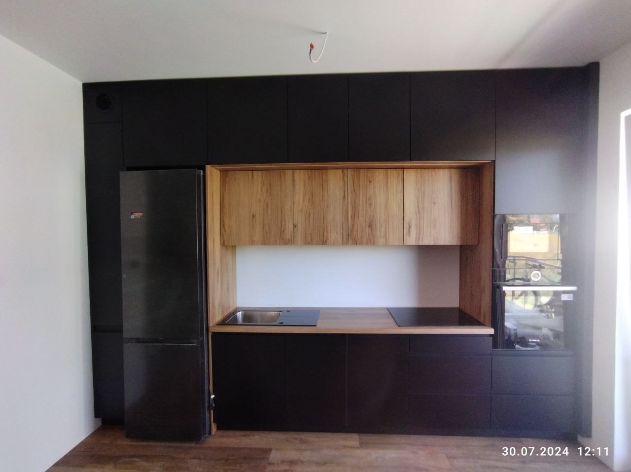 Nowoczesna kuchnia z czarnymi szafkami i drewnianymi akcentami, widoczny zlew, płyta indukcyjna i piekarnik w zabudowie, drewniana podłoga, surowy przewód elektryczny wystający z sufitu.