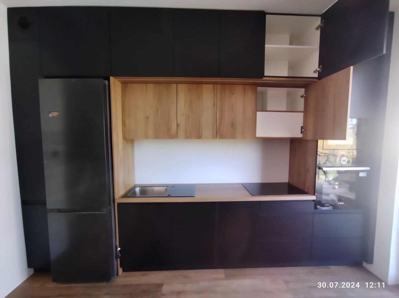 Nowoczesna kuchnia z zabudową w kolorze czarnym i drewnopodobnym, z otwartymi szafkami górnymi, widoczny zlew, płyta indukcyjna i lodówka.