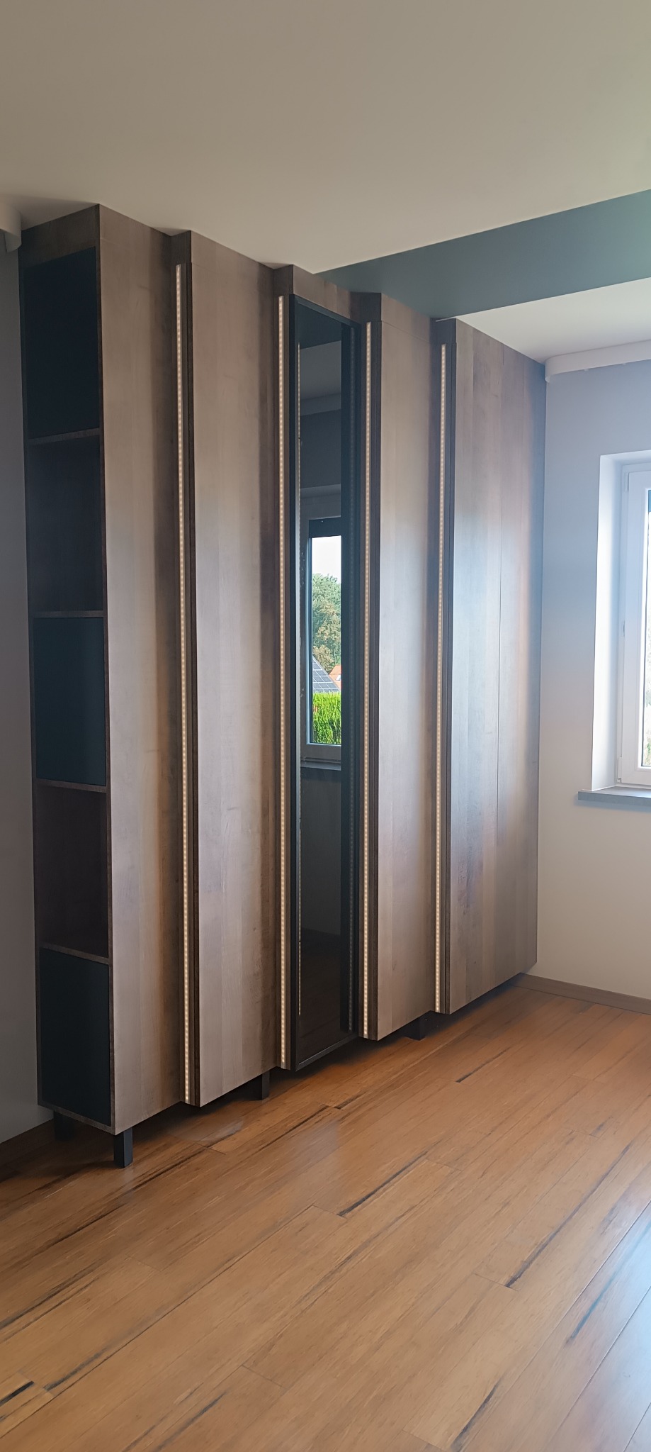 Wysoka, drewniana szafa z oświetleniem LED w pionowych listwach, z otwartą wnęką z lustrem, widok na zewnątrz przez okno.