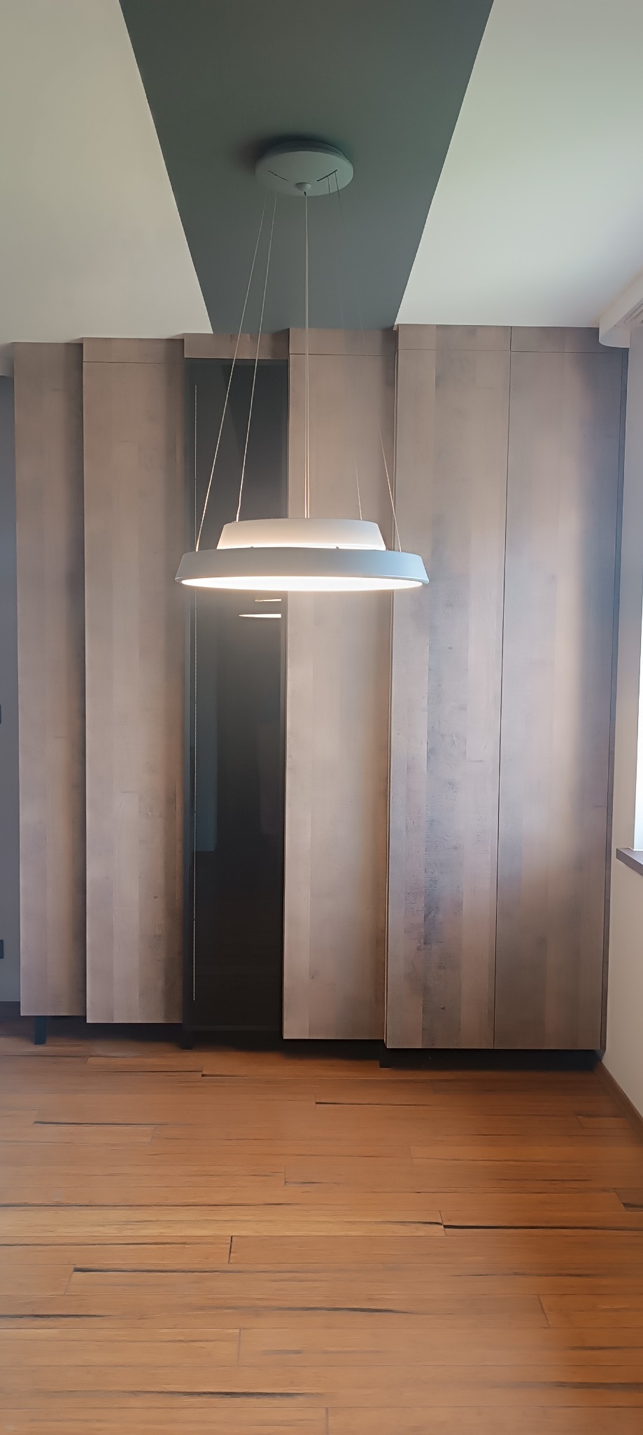 Wysoka zabudowa meblowa z drewnianymi frontami i czarnym, przeszklonym segmentem, oświetlona nowoczesną lampą wiszącą w minimalistycznym wnętrzu z drewnianą podłogą.