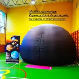 Rozłożone mobilne planetarium w sali gimnastycznej, obok baner reklamowy z grafiką planet i informacjami kontaktowymi, na podłodze różowa mata. Widoczne żółte ściany i okna.