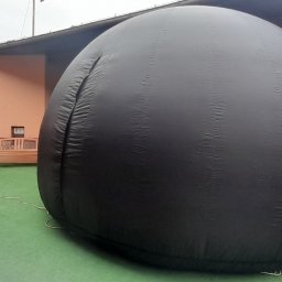 PLANETARIUM MOBILNE NATALIIA HARNYSTA - Czarny, dmuchany namiot sferyczny na zielonej powierzchni, obok placu zabaw dla dzieci i budynku.