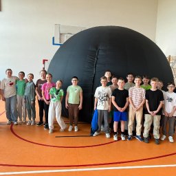 PLANETARIUM MOBILNE NATALIIA HARNYSTA - Grupa dzieci na tle rozłożonego, mobilnego planetarium w sali gimnastycznej. Uczniowie w różnym wieku, w codziennych ubraniach, stoją na tle pomarańczowej podłogi.