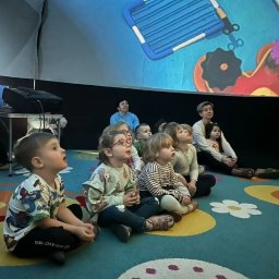 PLANETARIUM MOBILNE NATALIIA HARNYSTA - Grupa dzieci z opiekunami siedzi na podłodze z kolorowym wzorem, oglądając animowany film wyświetlany na ekranie. W tle widoczny sprzęt projekcyjny.