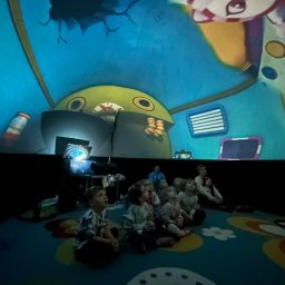 PLANETARIUM MOBILNE NATALIIA HARNYSTA - Grupa dzieci siedzi na podłodze, oglądając bajkę wyświetlaną na zakrzywionym ekranie. Widoczny projektor i fragmenty animacji z postaciami. Kolorowa podłoga.