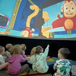 PLANETARIUM MOBILNE NATALIIA HARNYSTA - Grupa dzieci siedzi na podłodze, oglądając bajkę z małpką wyświetlaną na ekranie. Jedna dziewczynka wskazuje palcem na ekran. Sala zabaw z kolorowym dywanem.