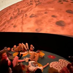 PLANETARIUM MOBILNE NATALIIA HARNYSTA - Grupa dzieci leżących na podłodze w planetarium, oglądających projekcję powierzchni Marsa na kopule. Kolorowy dywan z kwiatami dodaje radosnego akcentu.