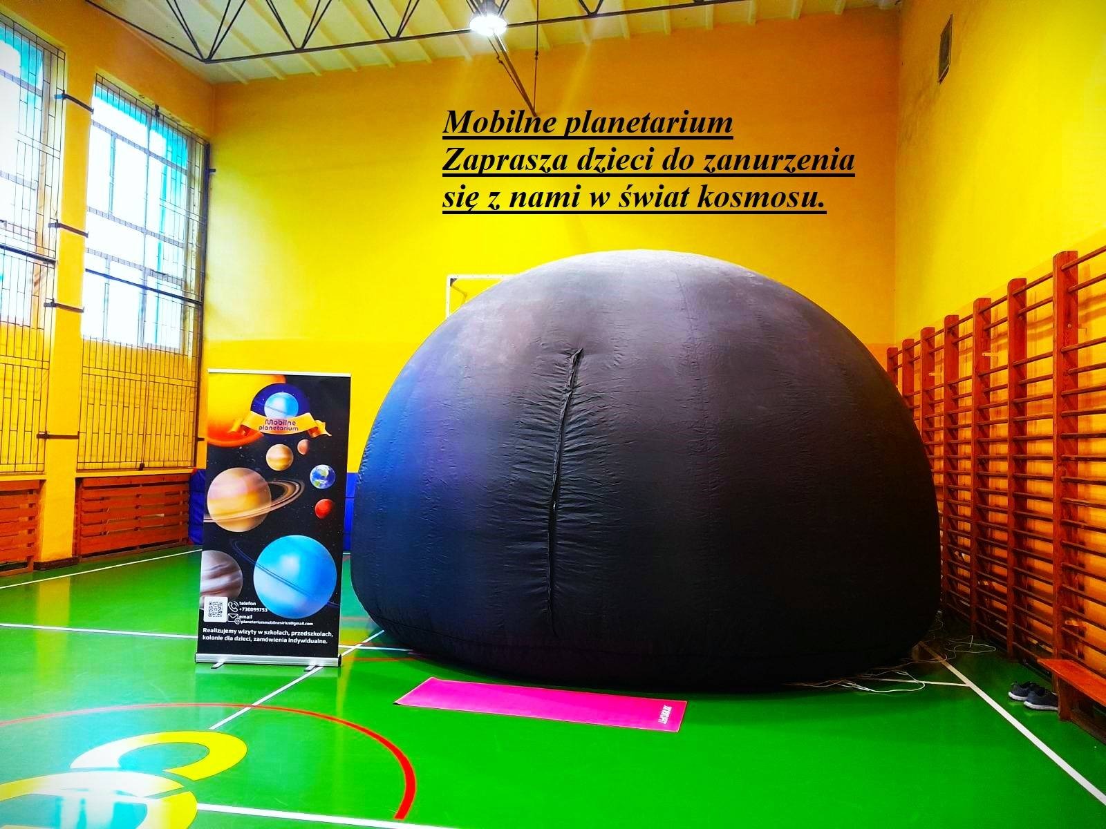 Rozłożone mobilne planetarium w sali gimnastycznej, obok baner reklamowy z grafiką planet i informacjami kontaktowymi, na podłodze różowa mata. Widoczne żółte ściany i okna.