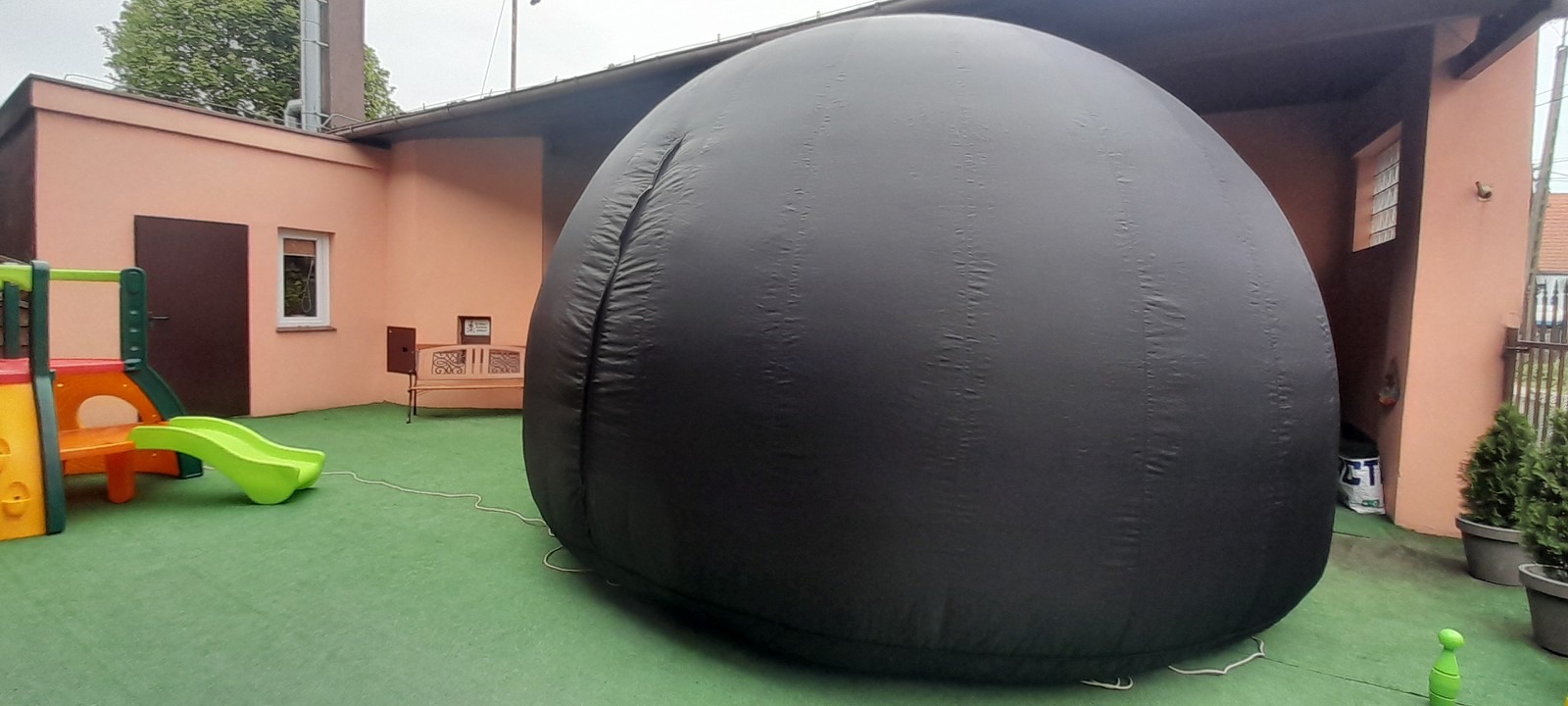 Czarny, dmuchany namiot sferyczny na zielonej powierzchni, obok placu zabaw dla dzieci i budynku.