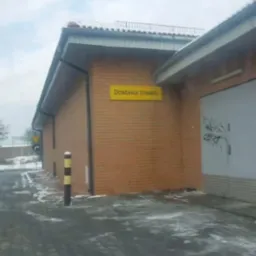 Budynek z napisem 'Dostawa towaru' na ceglanej ścianie, częściowo pokryty śniegiem, z zaszronionym chodnikiem i słupkiem ostrzegawczym w zimowy dzień.