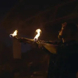 Mężczyzna z brodą wykonuje pokaz z pochodniami na tle ciemnej sceny z logo KA-BOOM Fireshow.