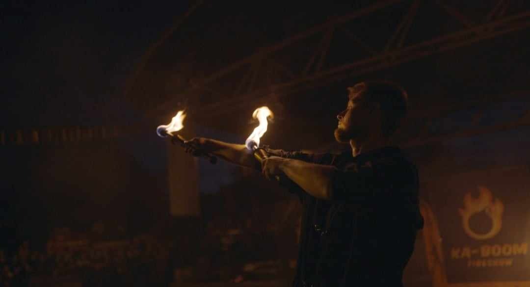 Mężczyzna z brodą wykonuje pokaz z pochodniami na tle ciemnej sceny z logo KA-BOOM Fireshow.