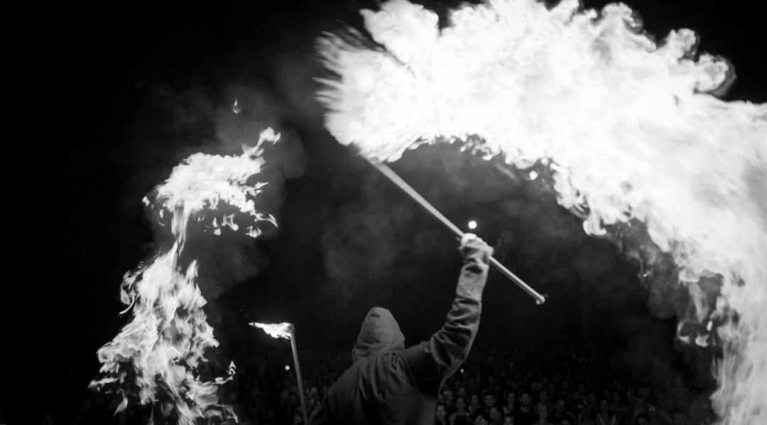 Czarno-białe zdjęcie osoby w kapturze, odwróconej tyłem, trzymającej płonące kije na tle publiczności, z dużymi kłębami ognia.