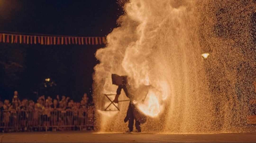 Uliczny pokaz ognia: osoba w ciemnym stroju manipuluje płonącymi rekwizytami na tle tłumu za barierkami i wiszących flagietek.