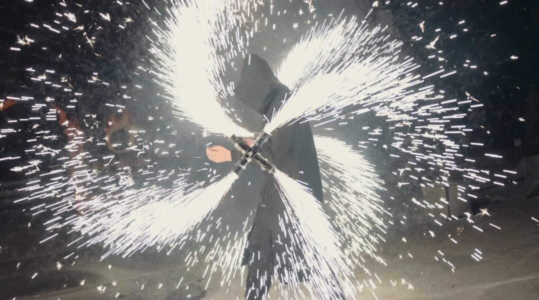 Osoba w kapturze trzymająca w skrzyżowanych rękach cztery palące się fontanny iskier, tworzące efektowny pirotechniczny pokaz.