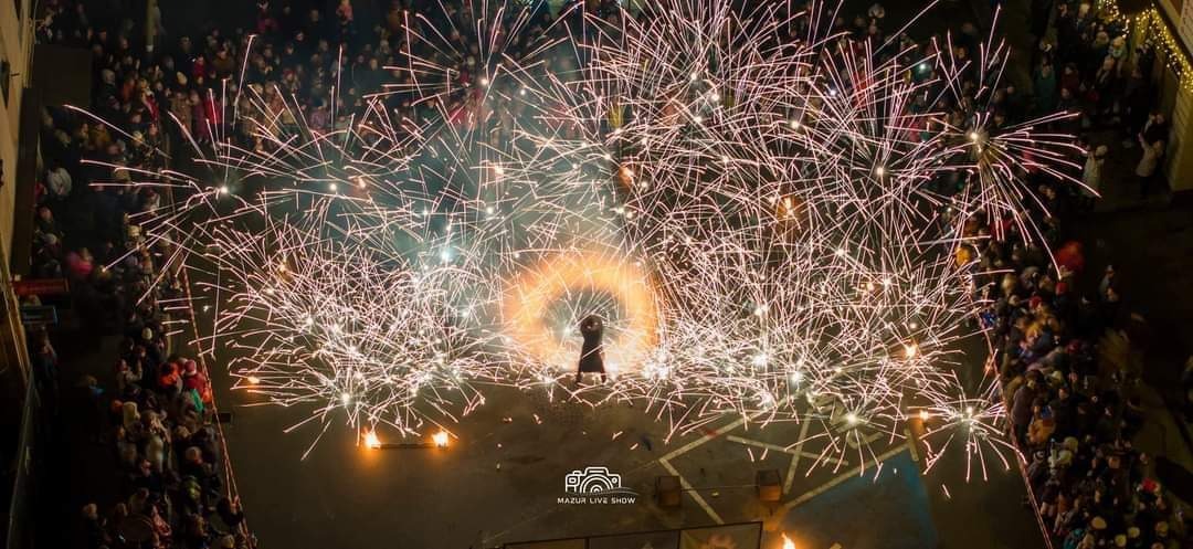 Widok z góry na pokaz sztucznych ogni na ulicy, z sylwetką osoby stojącej w płonącym okręgu i tłumem widzów w tle.