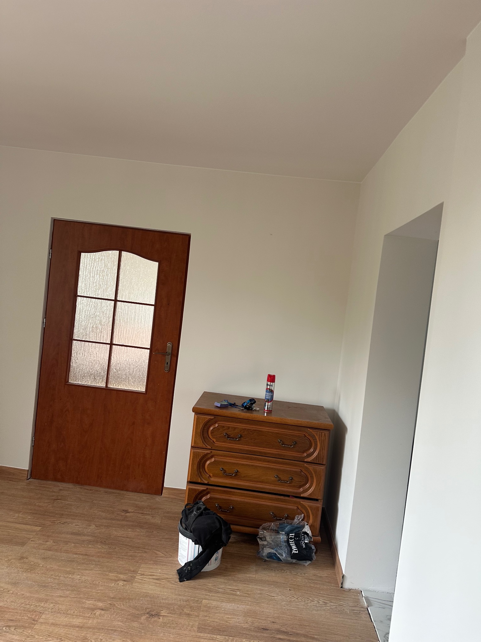 Jasne wnętrze po malowaniu: świeżo pomalowane ściany, drewniana komoda i drzwi z szybą. Widoczne akcesoria malarskie. Podłoga z paneli.