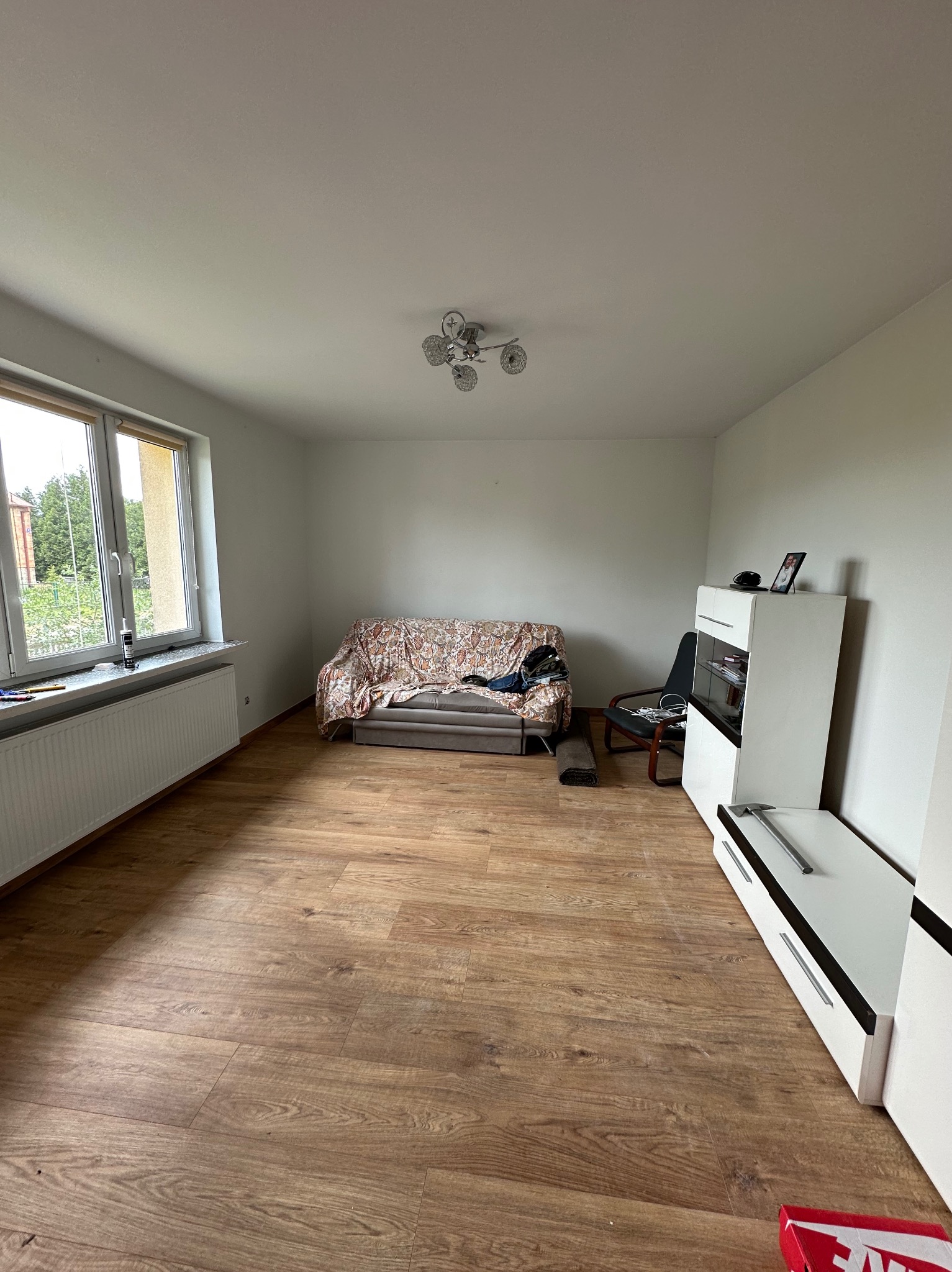 Jasne wnętrze pokoju z panelami podłogowymi, grzejnikiem pod oknem i meblami. Widoczny fragment sofy i szafki RTV. Na parapecie leżą narzędzia.