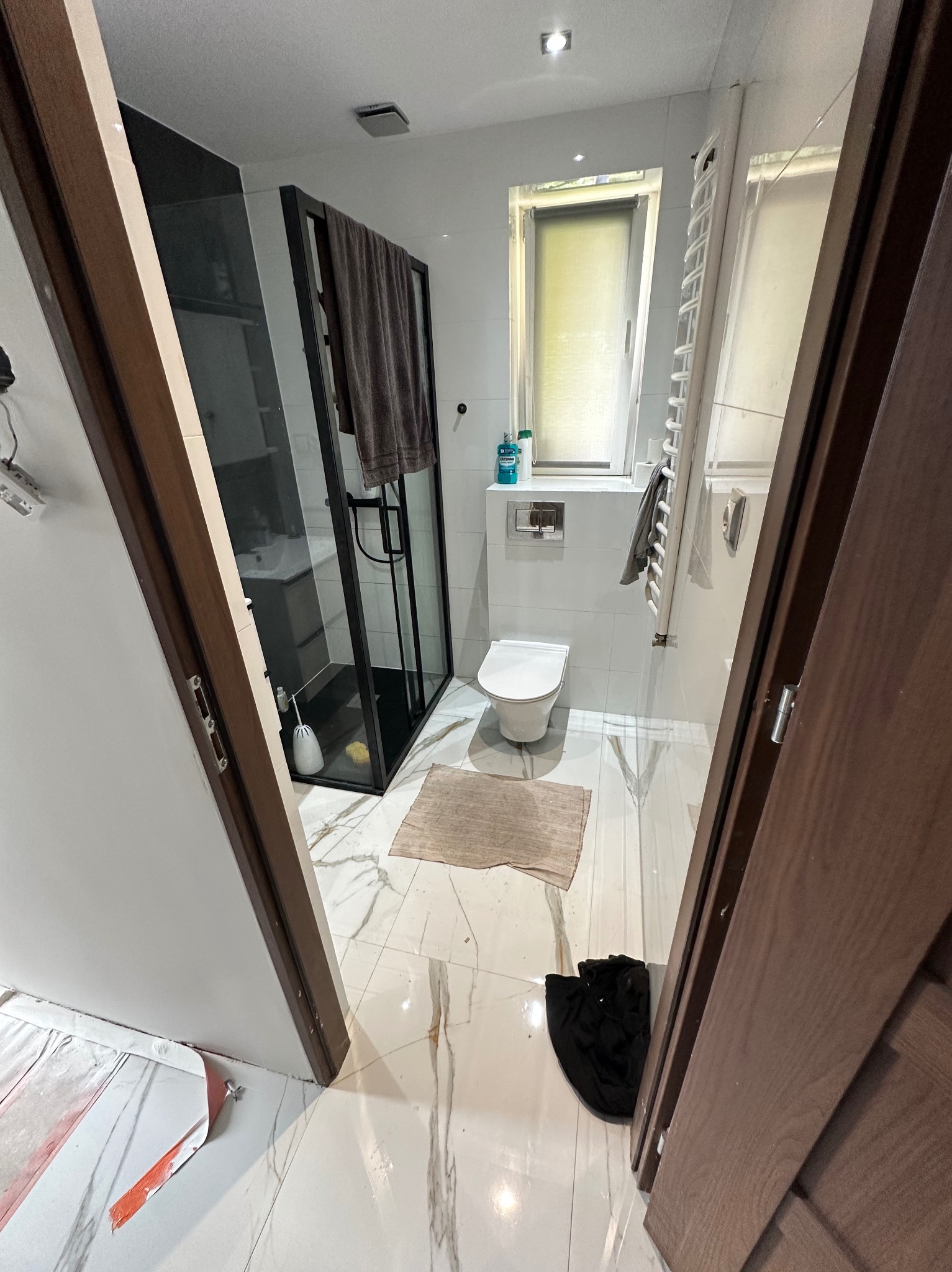 Wykończona łazienka z kabiną prysznicową typu walk-in, białą armaturą i marmurową posadzką, widziana zza otwartych, drewnianych drzwi. Częściowo widoczne zabezpieczenia podłogi.